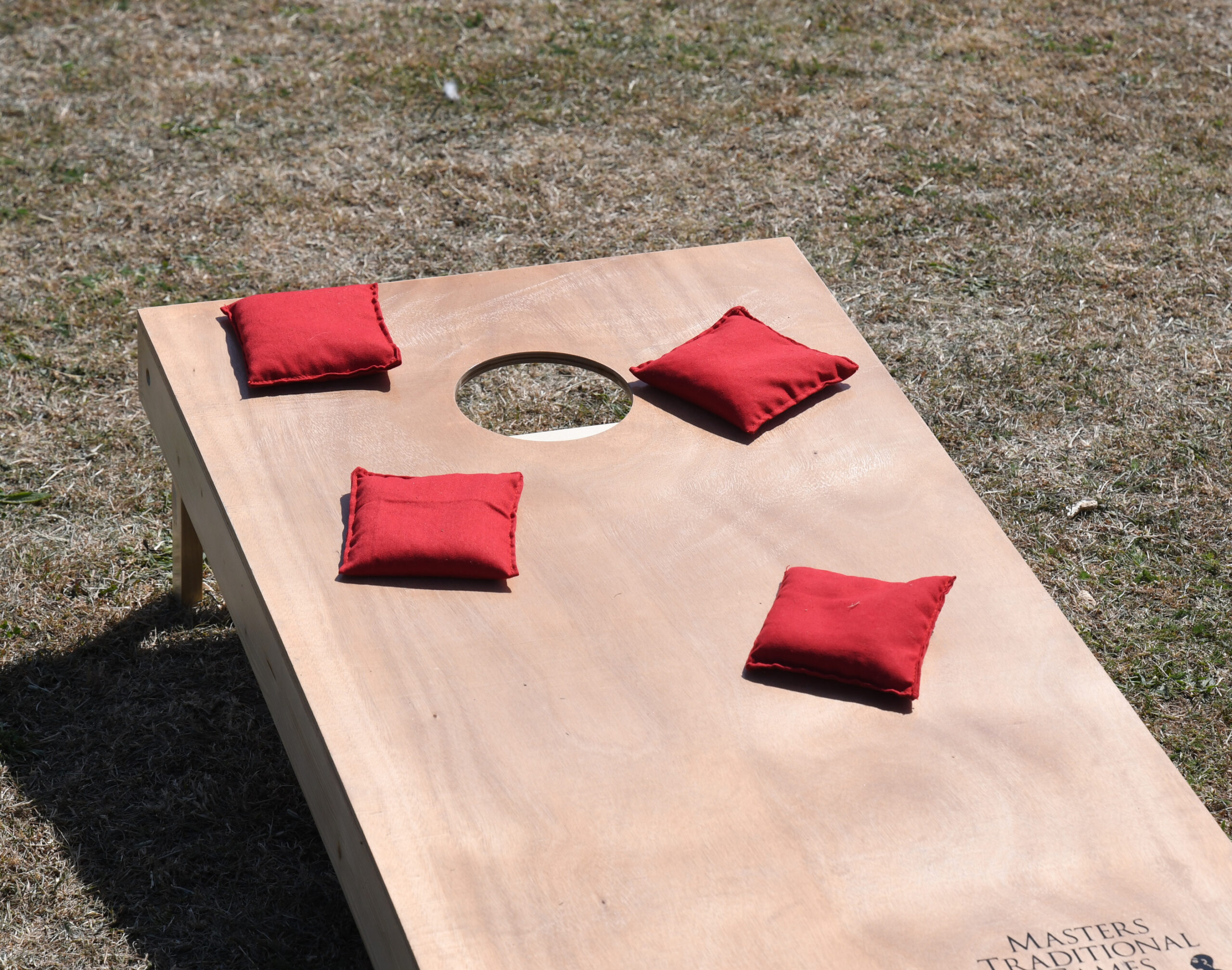 masters-cornhole-10 - Masters Traditional Games Outdoor wooden cornhole game board with a circular hole and red cushions placed on its surface, set on dry grass.