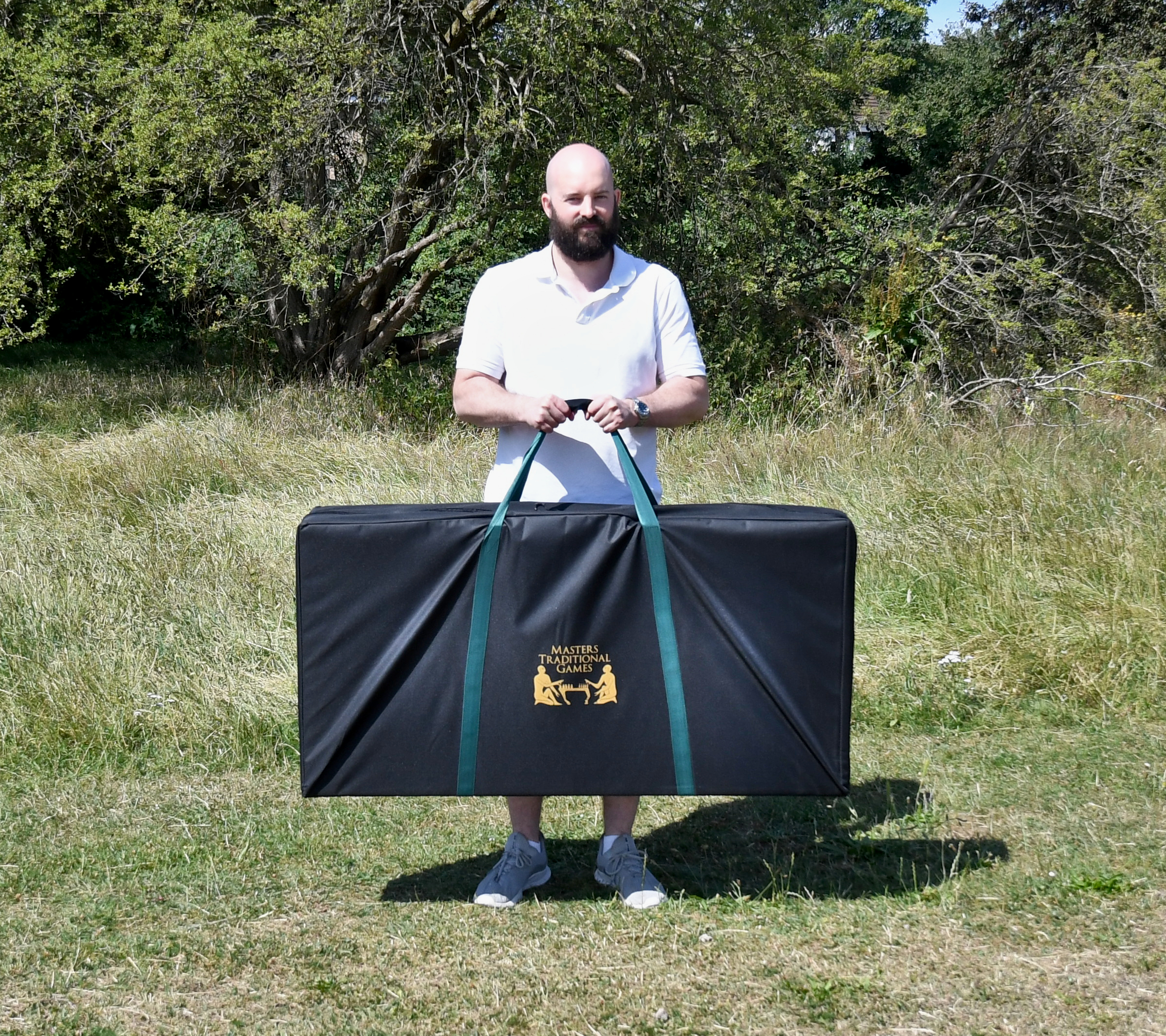 masters-cornhole-12 - Masters Traditional Games Bearded man in a white polo standing in a grassy field, holding a large black bag with green handles that reads Masters Traditional Games.