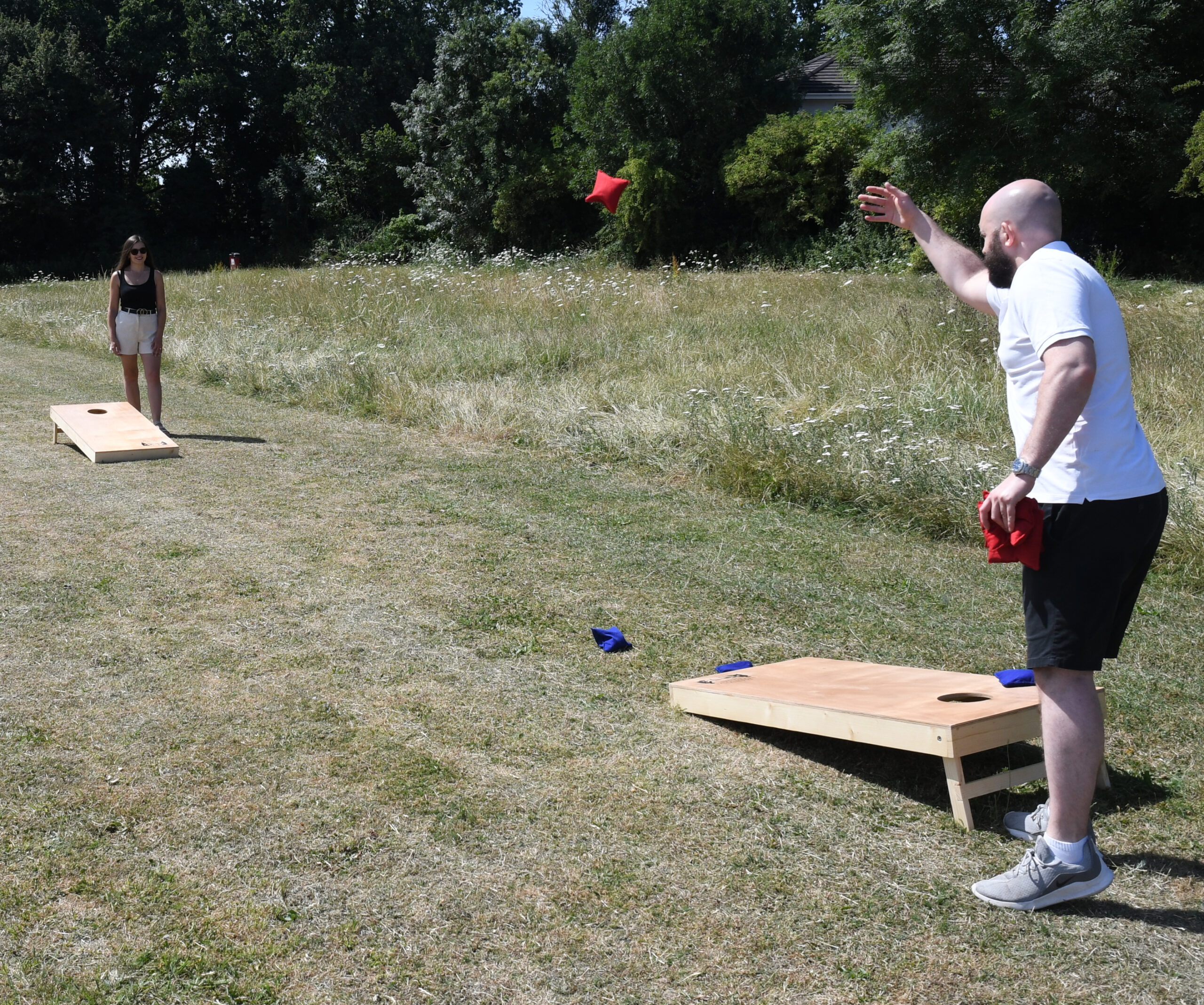 masters-cornhole-3 - Masters Traditional Games Man with a beard in a white shirt throws a red bean bag toward a cornhole board in a grassy park; woman in black top and white shorts watches in the background.