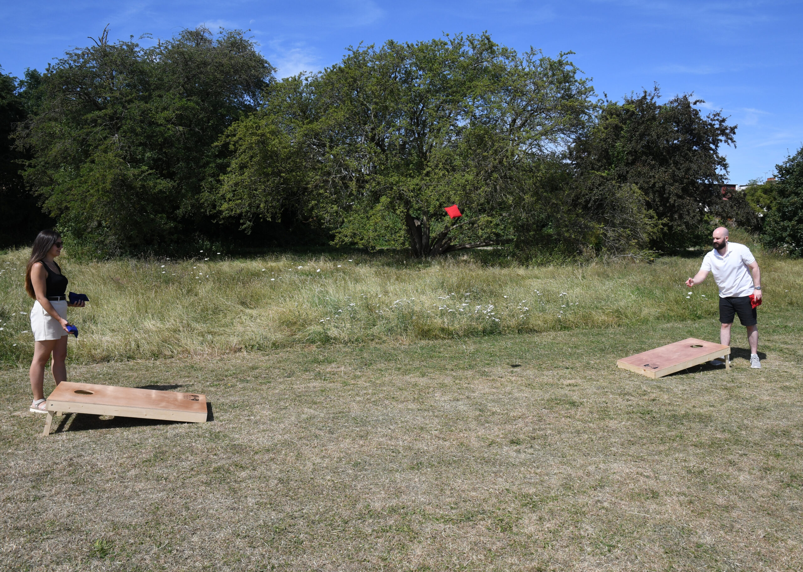 masters-cornhole-5 - Masters Traditional Games Two people play cornhole in a sunny field, a red bag flies toward a wooden board.