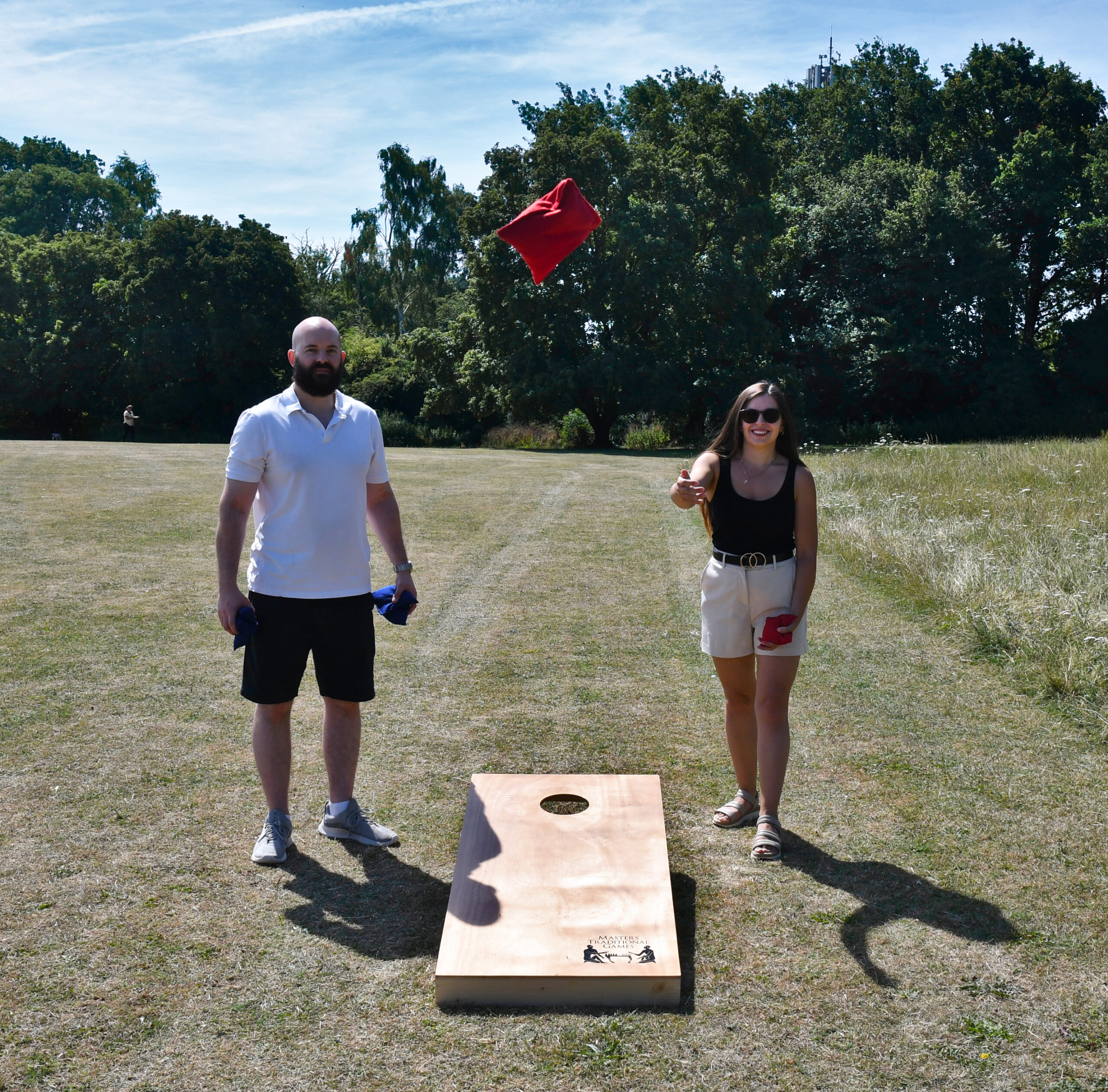 masters-cornhole-6 - Masters Traditional Games Two people stand on a sunny grassy field as a red bean bag flies toward a cornhole board nearby, in a park setting.