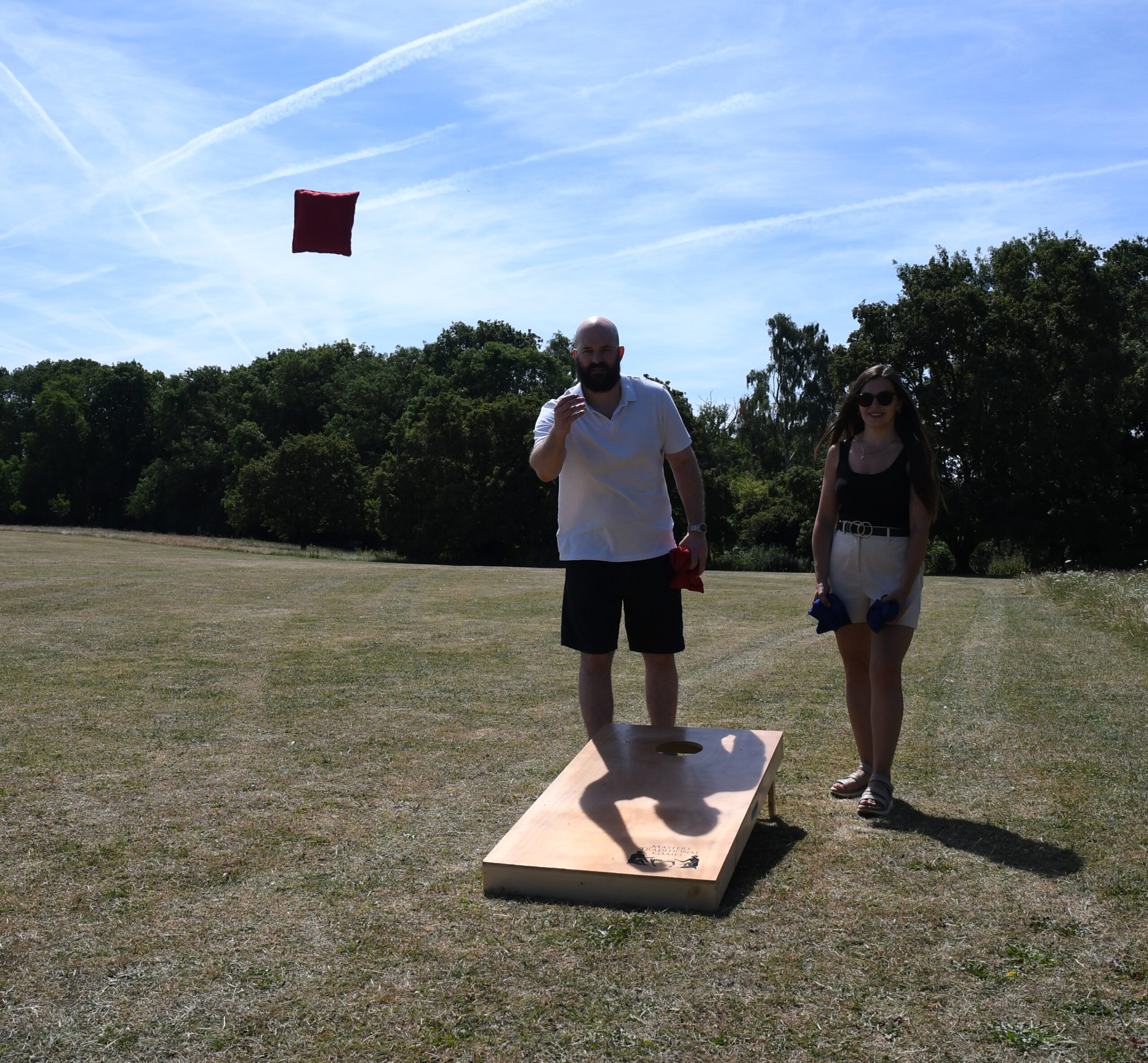 masters-cornhole-7 - Masters Traditional Games Man tossing a red bean bag toward a wooden cornhole board as a woman stands nearby outdoors on a sunny day with trees in the background.