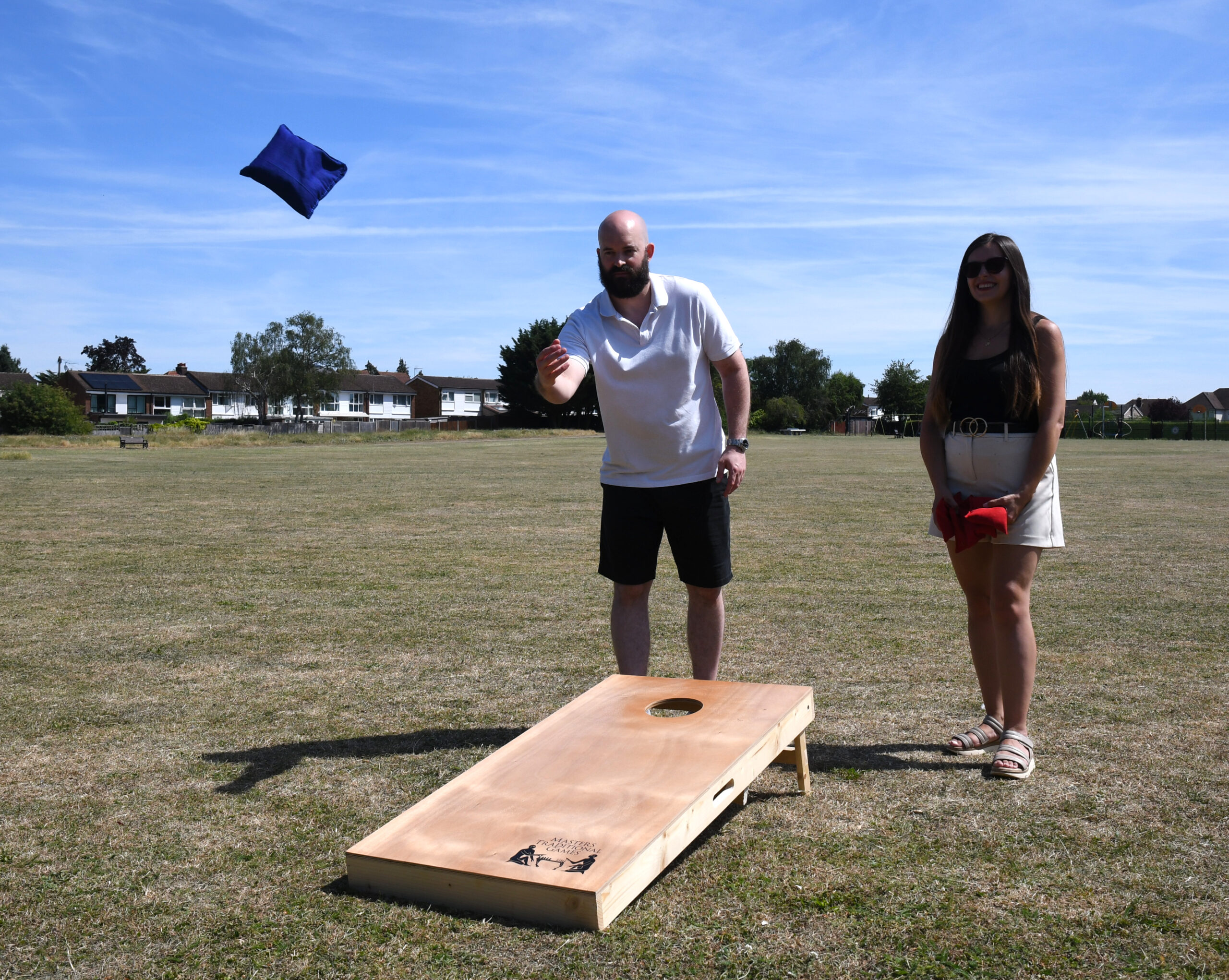 masters-cornhole-8 - Masters Traditional Games Man with a beard tosses a blue bean bag toward a cornhole board while a smiling woman watches on a sunny, open field.