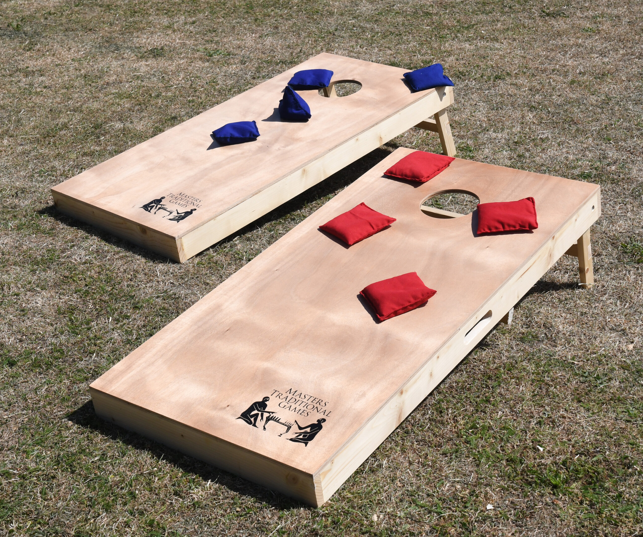 masters-cornhole-9 - Masters Traditional Games Wooden cornhole boards on dry grass with red and blue beanbags around the holes; outdoor lawn game setup.