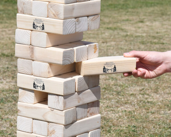 masters-giant-wooden-tumble-tower-10 - Masters Traditional Games Outdoor wooden Jenga tower being played, hand pulling a block from the stack; blocks show Masters Traditional Games logo.