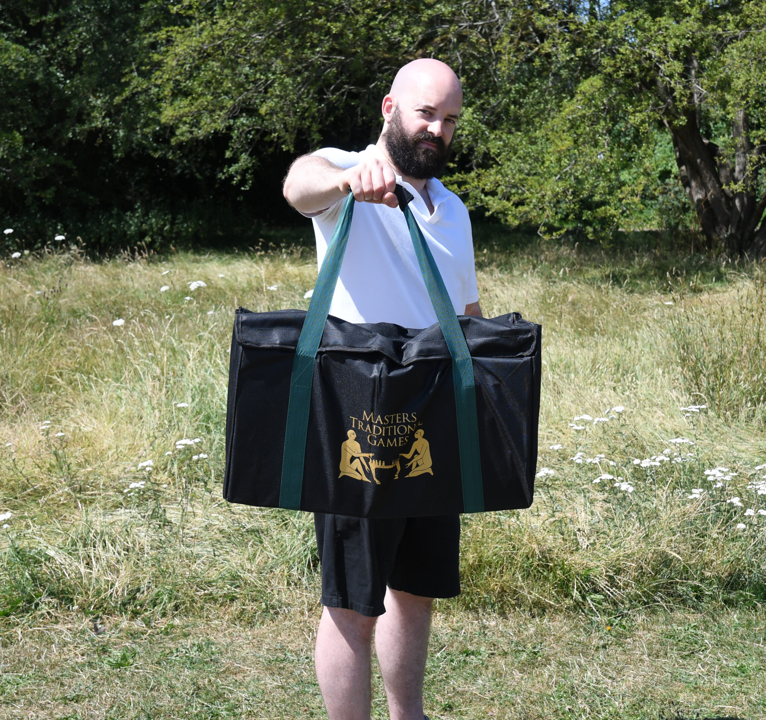 masters-giant-wooden-tumble-tower-12 - Masters Traditional Games Bearded bald man in a white shirt holding a large black bag with green straps in a grassy field.