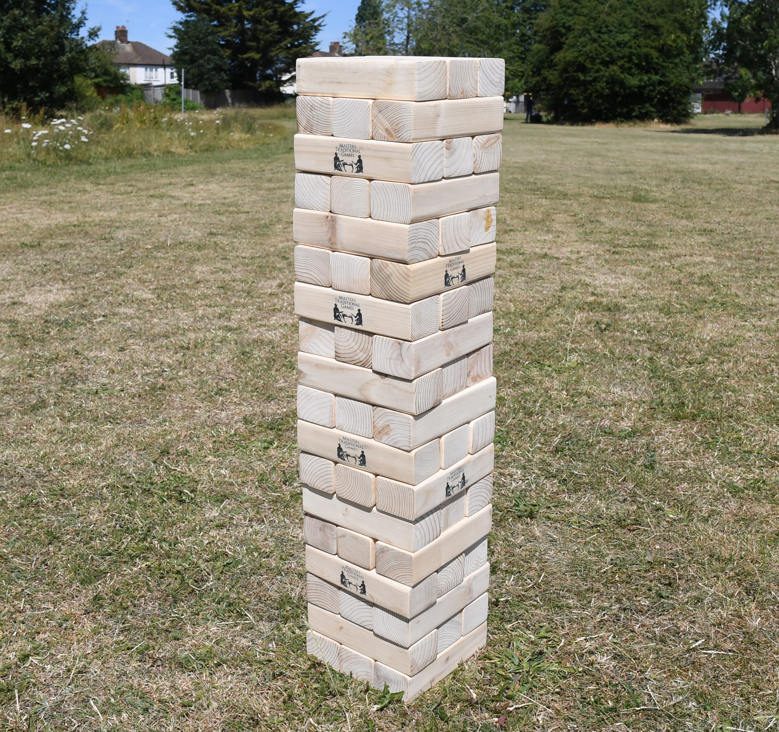masters-giant-wooden-tumble-tower-2 - Masters Traditional Games Tall wooden block tower standing on a grassy field with trees and houses in the background.