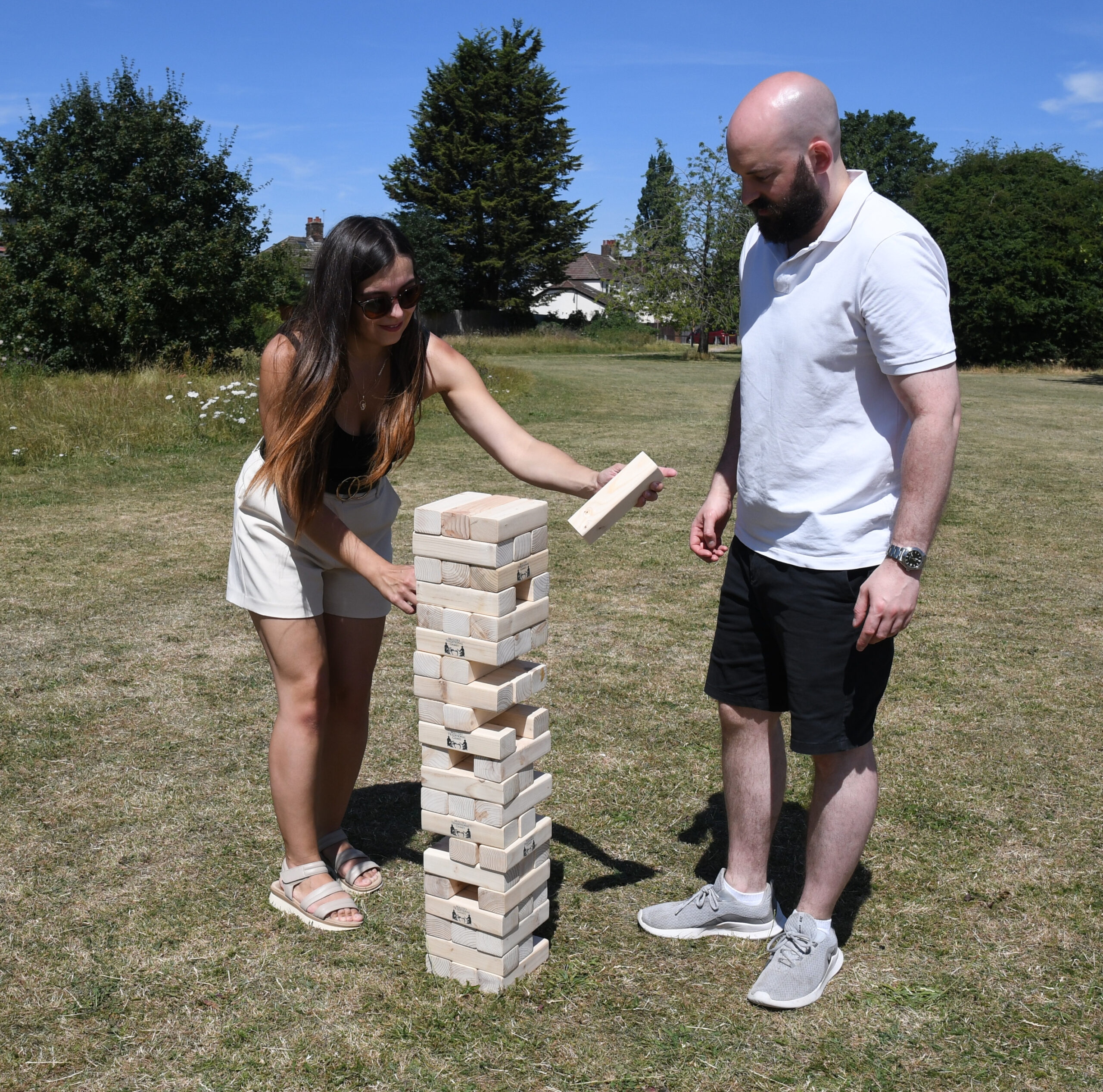 masters-giant-wooden-tumble-tower-3 - Masters Traditional Games Two people outdoors on a sunny day stack large wooden blocks for a giant Jenga game.