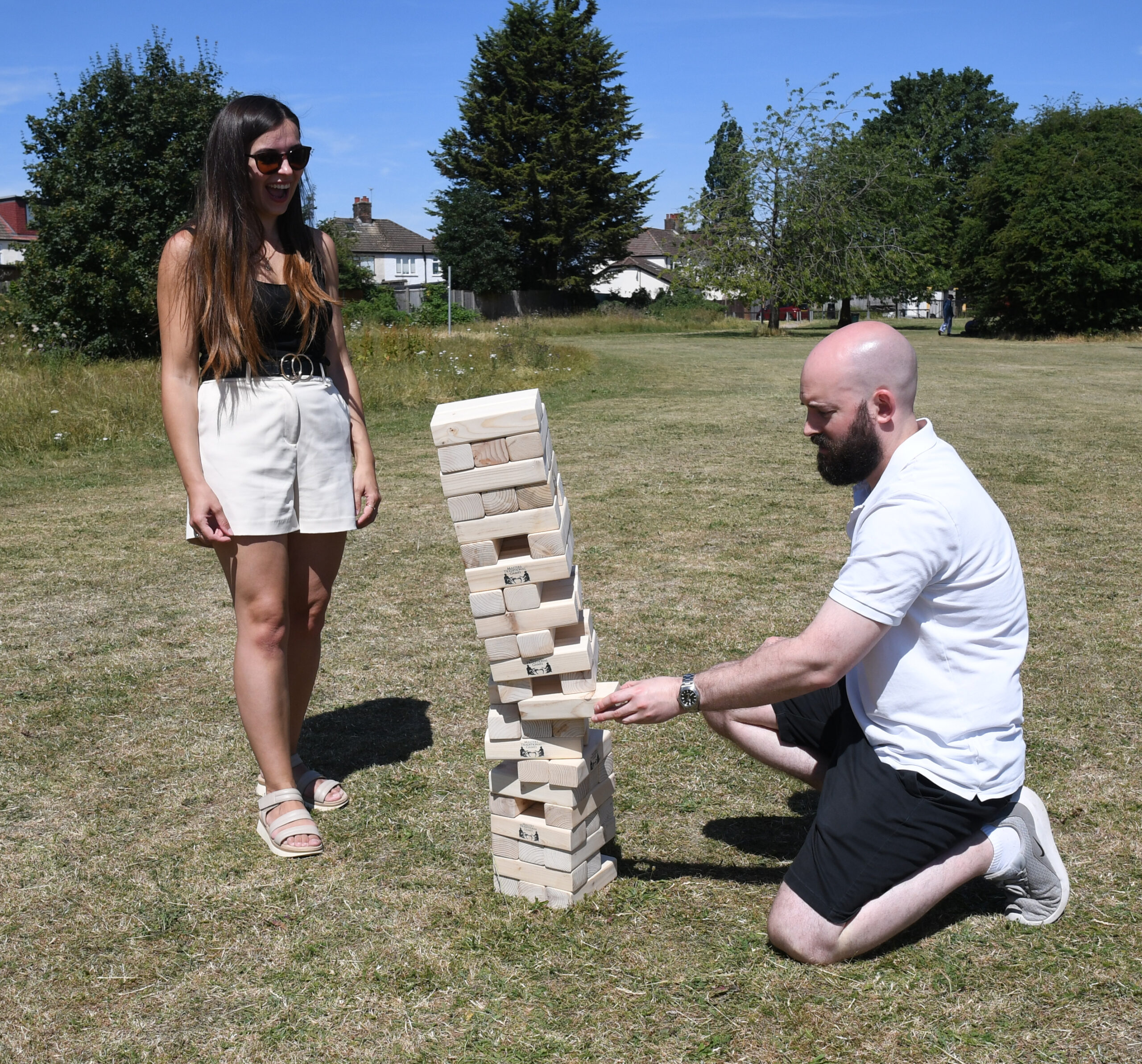 masters-giant-wooden-tumble-tower-5 - Masters Traditional Games Young woman wearing sunglasses stands outdoors beside a tall wooden Jenga tower, as a man kneels nearby.