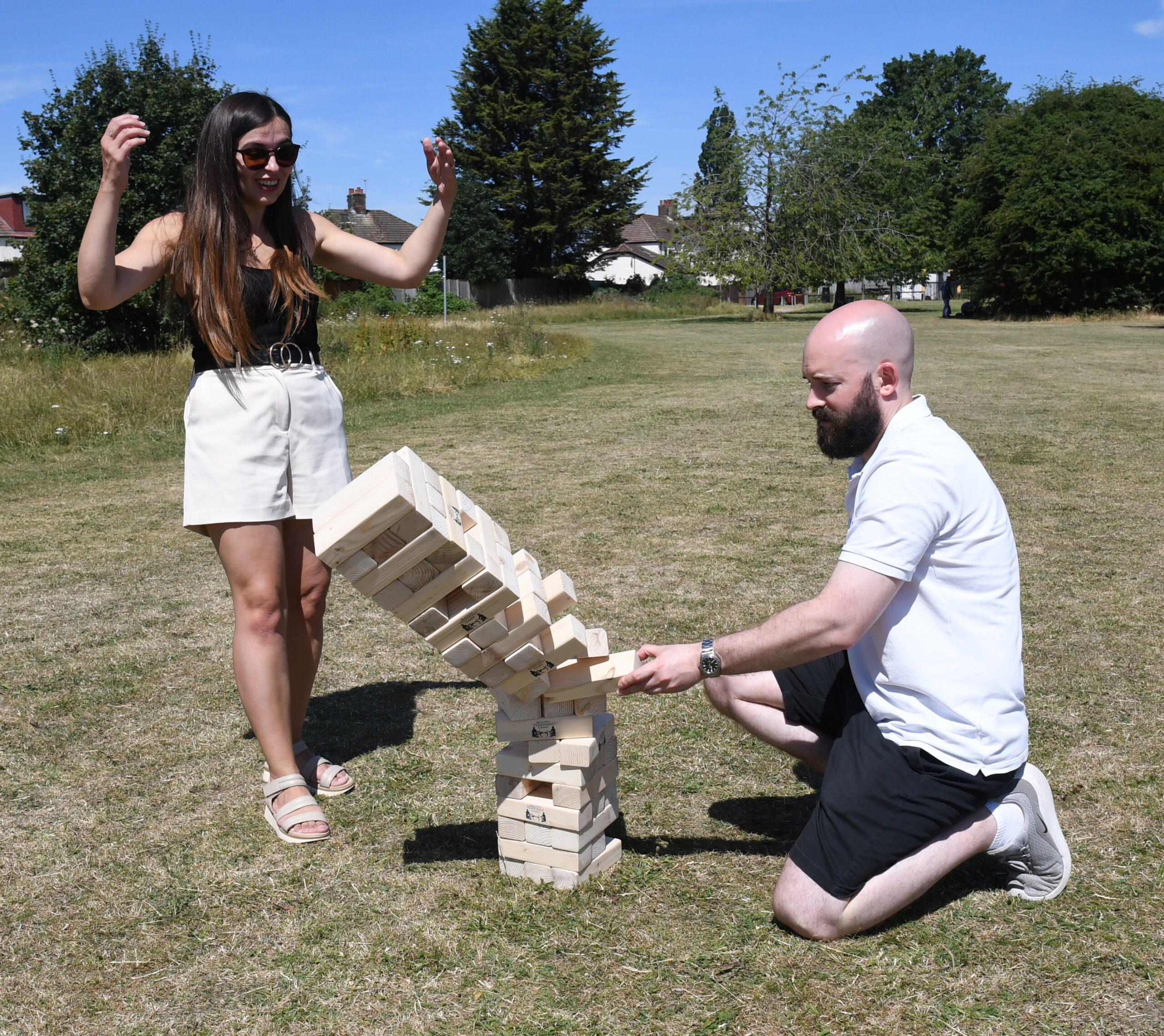 masters-giant-wooden-tumble-tower-6 - Masters Traditional Games Two adults in a park play giant Jenga, with a woman standing and smiling as a wooden block tower leans.