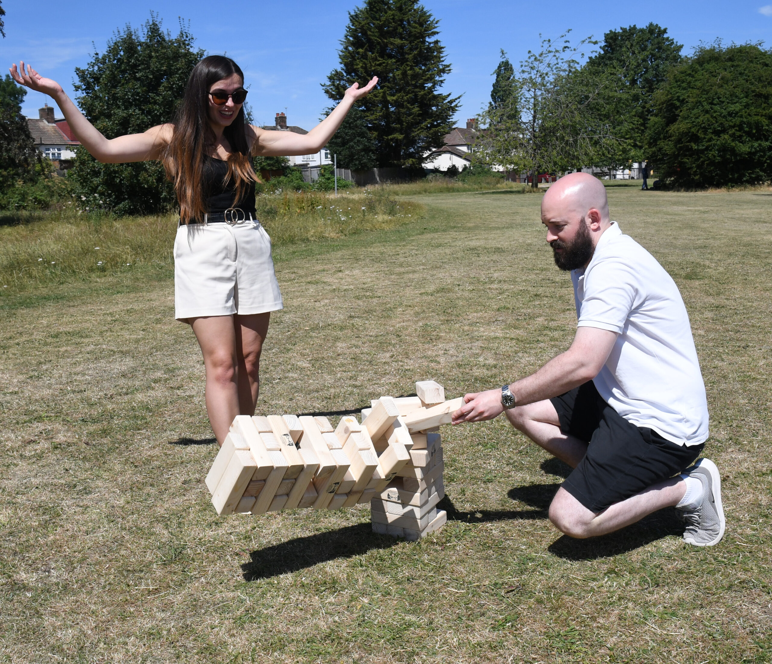 masters-giant-wooden-tumble-tower-7 - Masters Traditional Games Two people outdoors playing a giant wooden Jenga tower in a sunny park; woman with arms raised and sunglasses stands nearby as the man kneels to place blocks.