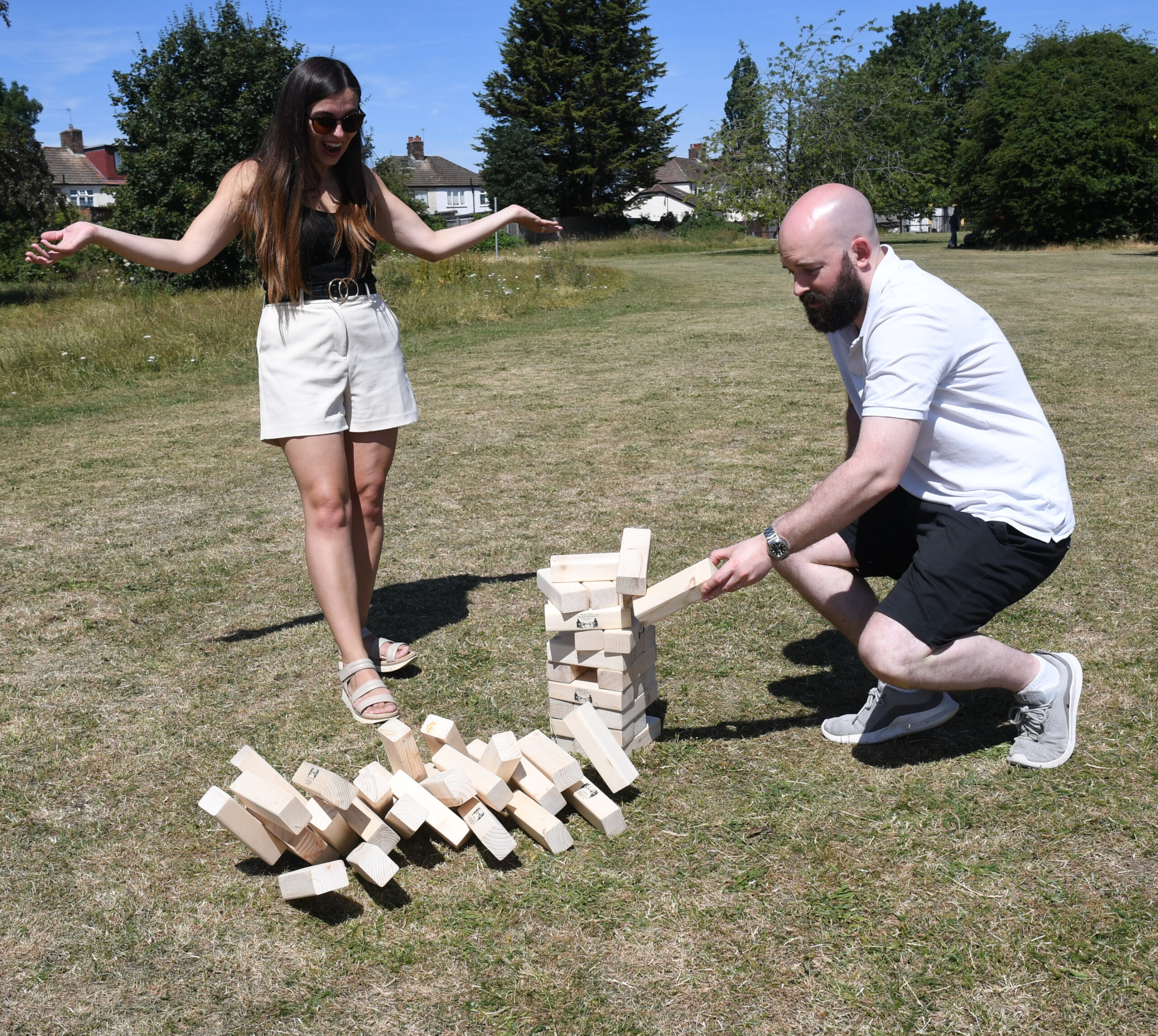 masters-giant-wooden-tumble-tower-8 - Masters Traditional Games Two adults playing a large outdoor Jenga game in a sunny park; woman stands with arms out, man kneels to place blocks.
