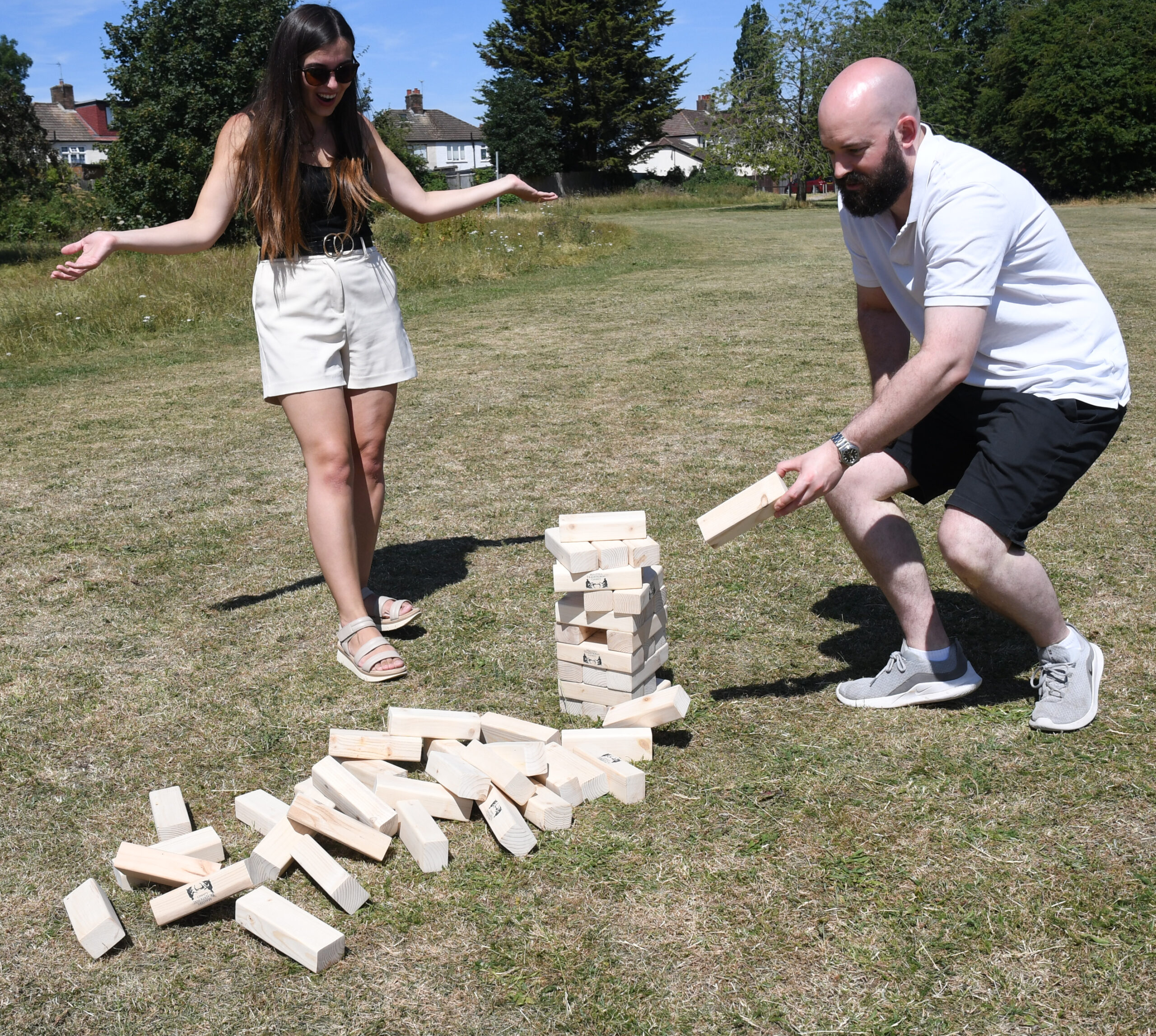 masters-giant-wooden-tumble-tower-9 - Masters Traditional Games Two people playing outdoor giant Jenga on a sunny lawn; a woman in white shorts and sandals gestures while a bearded man in a white polo stacks wooden blocks.