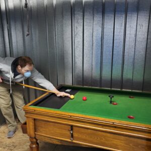 bar-billiards-jelks-losangeles-1 - Masters Traditional Games - Masters Traditional Games Man wearing a face mask leans over a pool table, aiming with a cue at a yellow ball while red balls sit on the green felt.