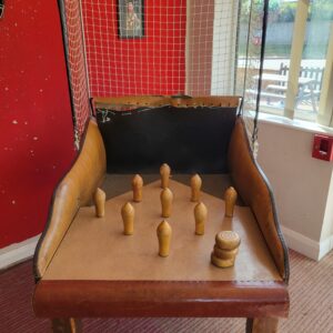 Wooden bowling pins arranged on a curved arcade table in front of a netted goal and red wall inside a game room.