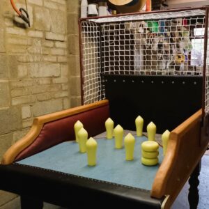 Small vintage arcade table with yellow game pegs on a blue playing surface, in a workshop area with brick wall and mesh enclosure behind.