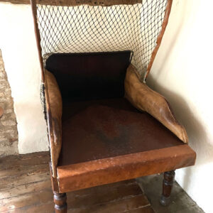 Old brown leather armchair with a metal mesh canopy in a rustic room, showing worn cushions and legs on a wooden floor.