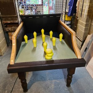 Tabletop arcade bowling game with yellow pins set up inside a wooden-framed play table; a stack of yellow discs sits at the front. Net barrier behind the pins.