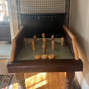 Vintage wooden tabletop bowling game with a green playing surface, pins arranged to be knocked over, enclosed by padded rails and a netted back panel in a living room setting.