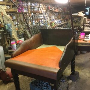 Brown leather bench seat with an orange cushion under a metal wire canopy in a cluttered workshop, with tools and shelves in the background.